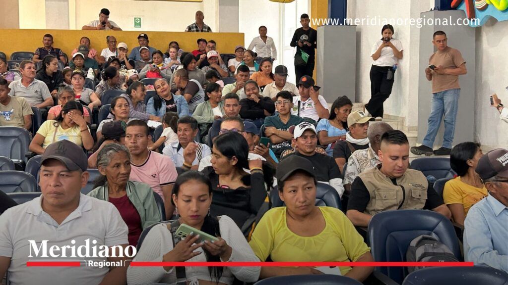 Auditorio en Caloto lleno de hombres y mujeres del norte del Cauca, sentados y atentos durante la audiencia pública por la paz. Algunos toman fotos o revisan sus celulares; decoraciones coloridas cuelgan en las paredes.