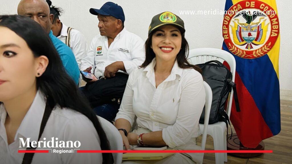 Maribel Perafán, secretaria de Gobierno del Cauca, sonríe sentada en una silla plástica blanca, vestida con camisa blanca y gorra institucional. Al fondo se observa la bandera de Colombia y otros asistentes a la audiencia pública en Caloto.