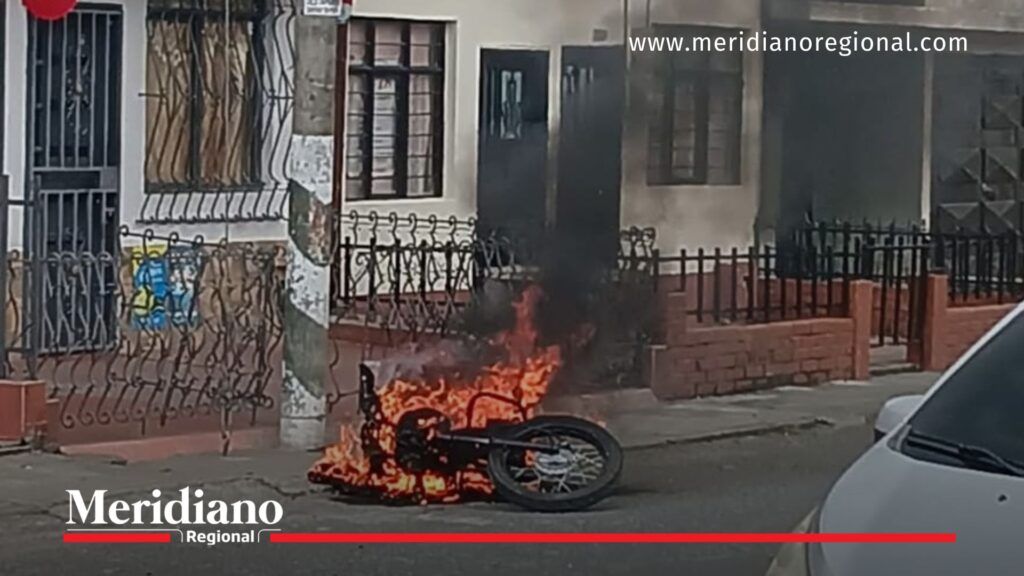 Motocicleta caída e incendiada frente a una vivienda en el barrio El Libertador de Popayán, con llamas intensas y humo negro, en una calle residencial con rejas y fachadas tradicionales