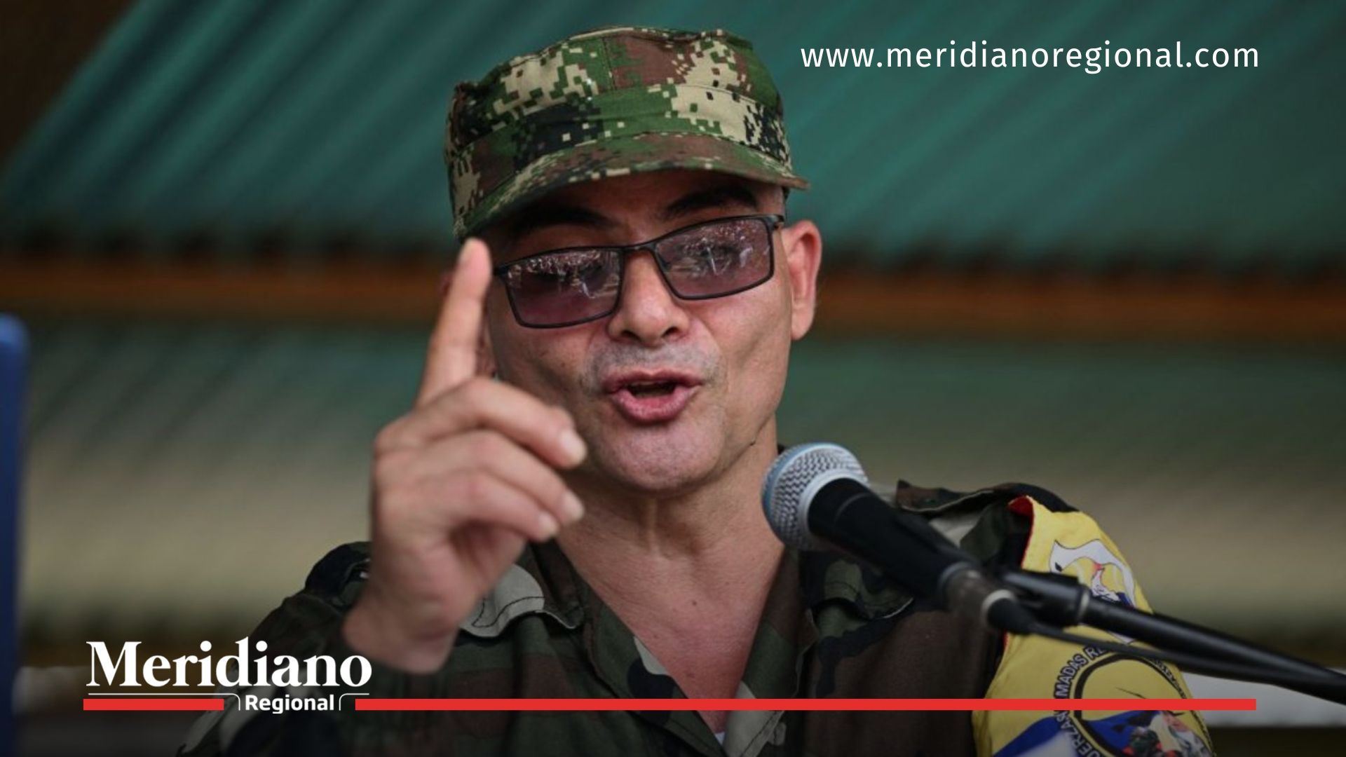Hombre con uniforme camuflado y gafas hablando frente a un micrófono en un campamento armado.