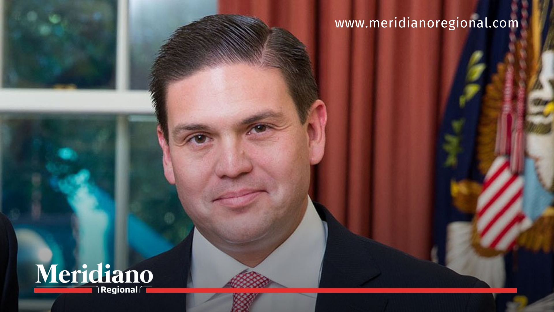 Hombre en un evento institucional, en un escenario formal con banderas y cortinas oficiales.
