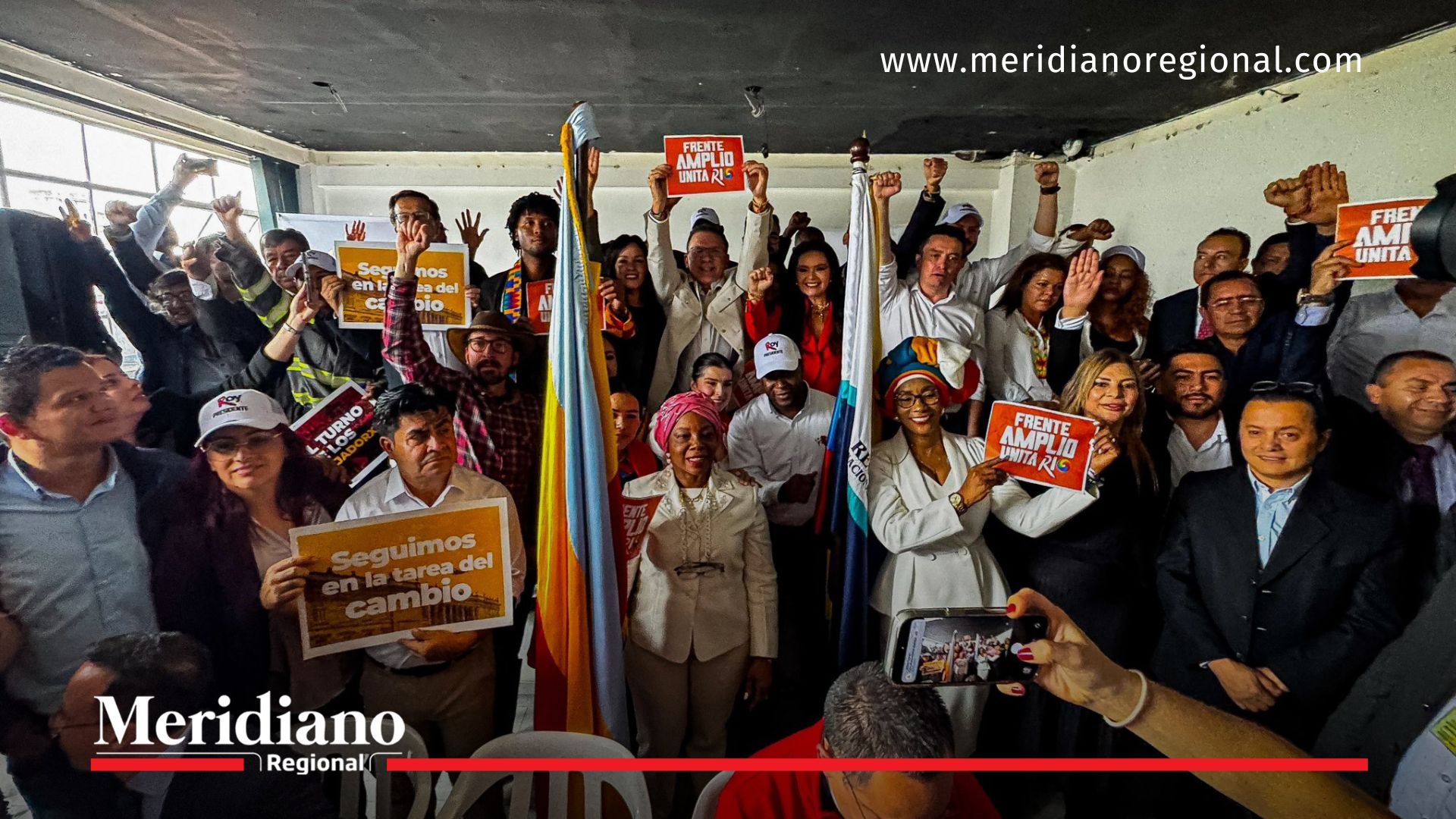 Grupo numeroso celebrando la presentación de la lista del Frente Amplio Unitario, con carteles, banderas y asistentes reunidos en un espacio interior.