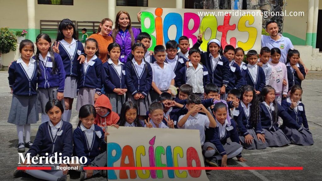 La imagen muestra a un grupo numeroso de estudiantes de primaria posando en el patio de la Institución Educativa Técnico Agropecuario Margarita Legarda, en la vereda Santa Leticia, Puracé. Los niños y niñas visten uniforme escolar azul y gris, y detrás de ellos se observan dos carteles elaborados por la comunidad estudiantil: uno que dice “Biobots” en letras grandes y coloridas, y otro que dice “Pacífico”, también decorado con colores vivos. Docentes y acompañantes se ubican detrás del grupo, sonriendo. El ambiente es alegre y refleja el entusiasmo por la jornada pedagógica del proyecto Biobots.