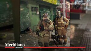 Firefighters in full turnout gear walk through a damaged street with green storefronts and debris on the ground, responding to an incident.