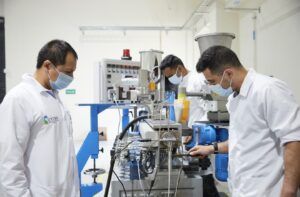 Three technicians in white coats and masks monitor a complex industrial machine in a clean lab setting.