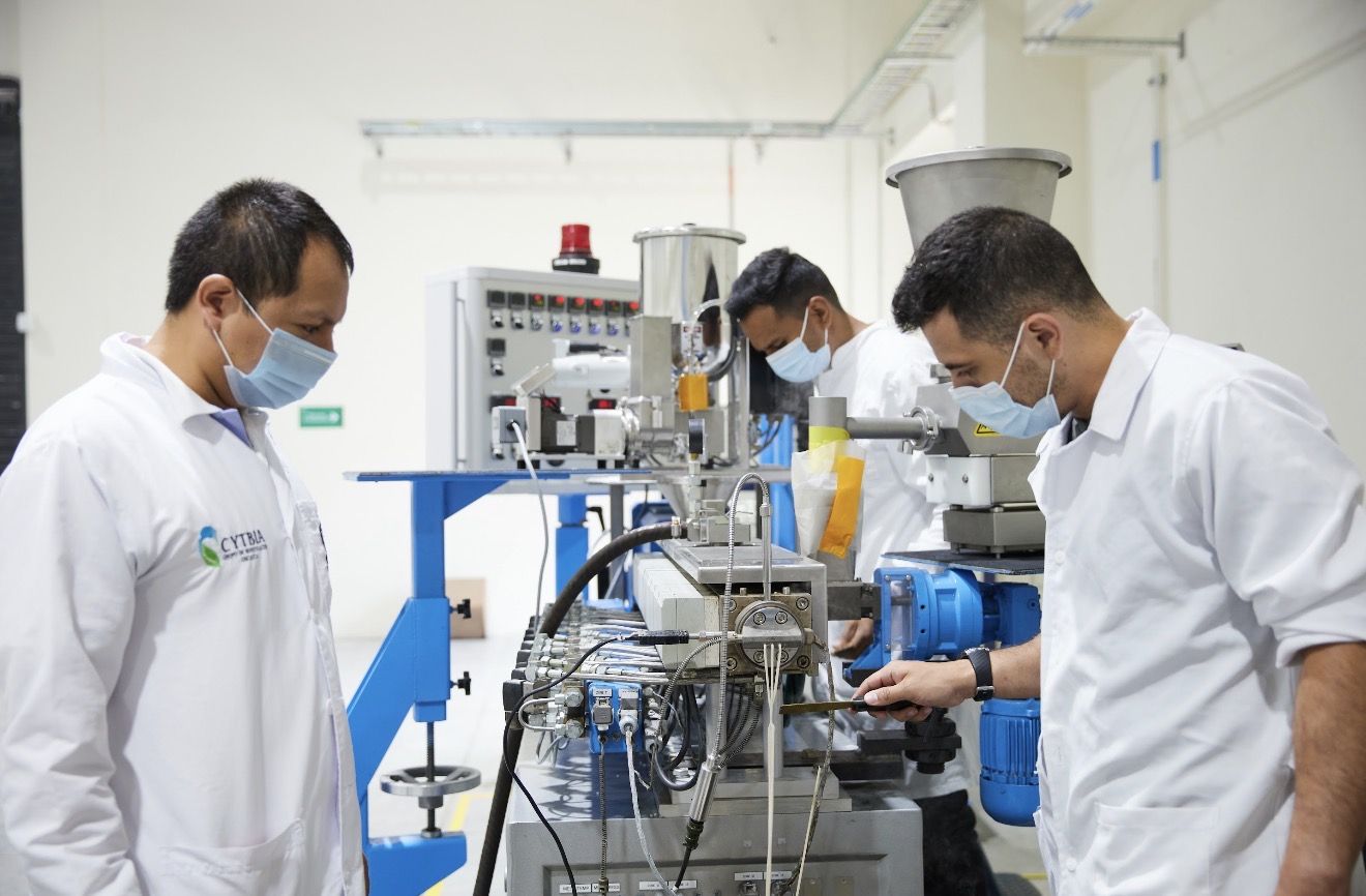 Three technicians in white coats and masks monitor a complex industrial machine in a clean lab setting.