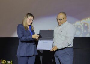 Woman in a blue suit hands a certificate to a man in a white shirt on stage, with flowers in the foreground.