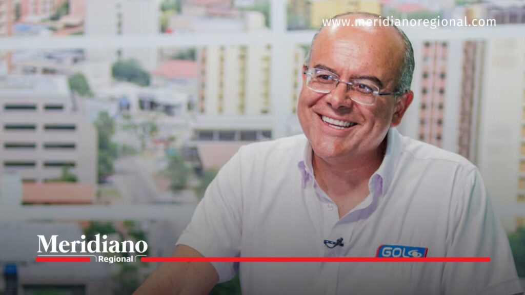 Ricardo Orrego sonríe durante una entrevista, vestido con una camisa blanca de Gol Caracol y gafas, con edificios desenfocados al fondo.