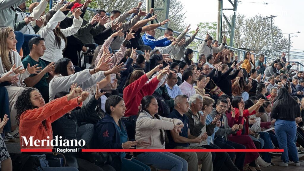 Padres y madres de familia sentados en graderías participan en una actividad grupal con las manos levantadas.