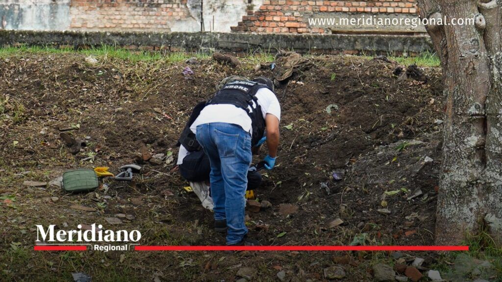 Investigador con chaleco y guantes azules inspecciona un terreno junto a un árbol, en el sector del atentado contra policías en Popayán.