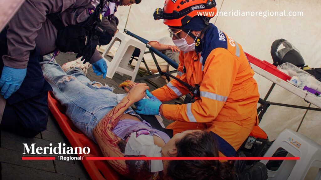Rescatista con uniforme naranja y guantes azules atiende a una persona con heridas simuladas sobre una camilla durante un ejercicio de emergencias.