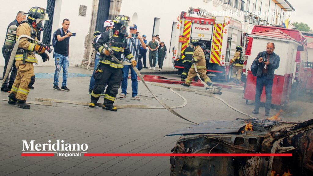 Bomberos de Popayán con equipos de protección y mangueras atienden un incendio controlado en un vehículo durante un ejercicio de emergencia.