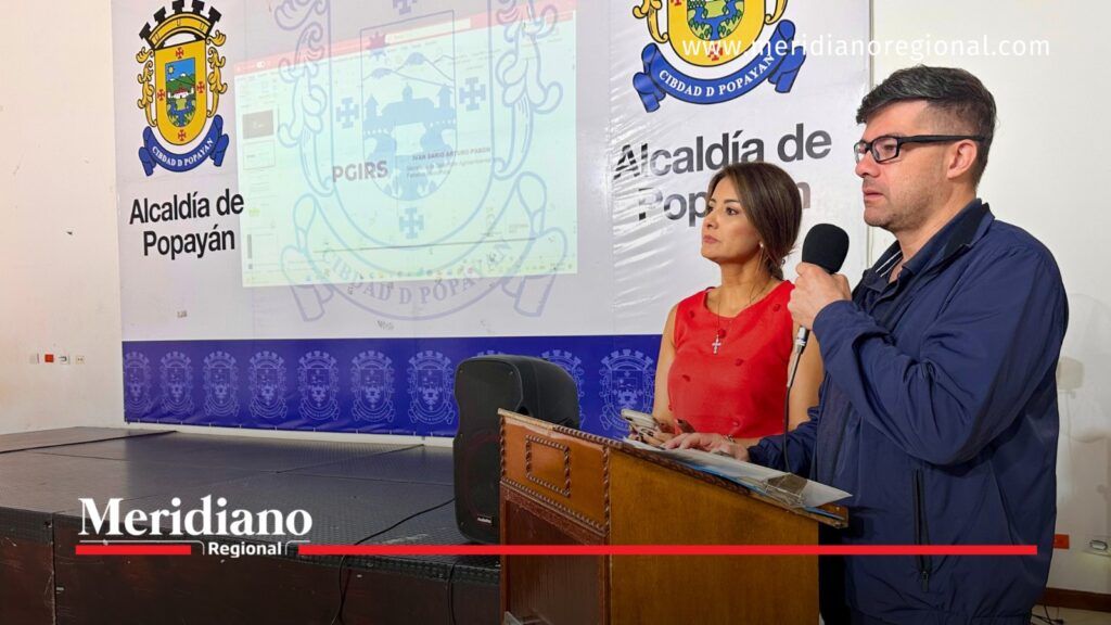 Hombre con chaqueta azul interviene con un micrófono junto a una mujer de rojo, frente a una presentación del PGIRS y los emblemas de la Alcaldía de Popayán.