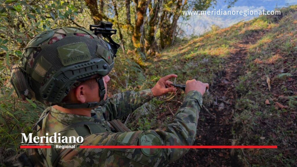 Militar del Ejército Nacional con uniforme camuflado y casco manipula un dispositivo junto a un camino rural rodeado de árboles y vegetación.