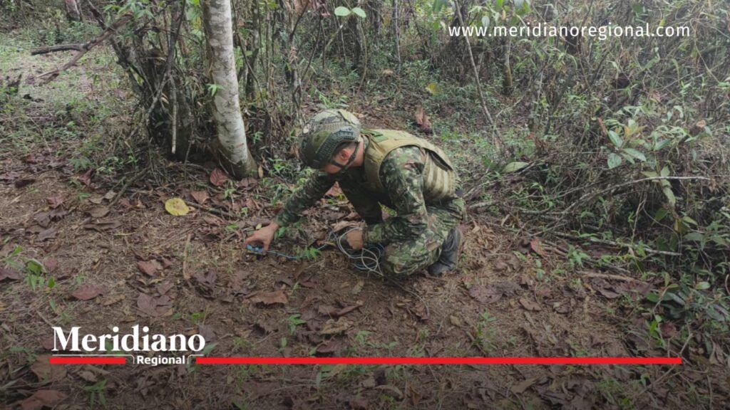 Militar con uniforme camuflado y equipo de protección inspecciona un elemento con cables sobre el suelo de una zona boscosa.